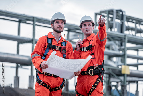 Engineer survey team wear uniform and helmet stand workplace checking blueprint project , radio communication and engineer box inspection work construction site with oil refinery background.