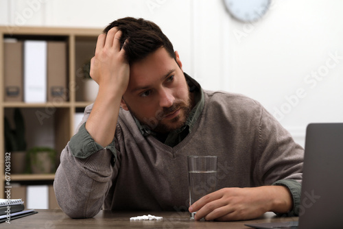Depressed man with glass of water and antidepressant pills at table indoors