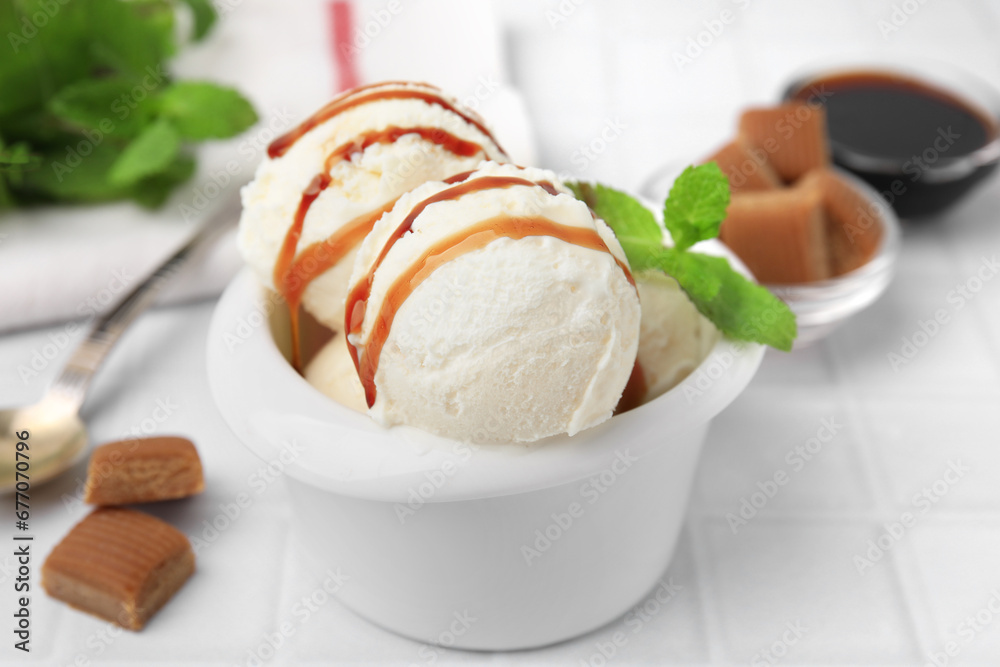 Scoops of tasty ice cream with mint leaves, candies and caramel sauce on white tiled table, closeup Scoops of tasty ice cream with mint leaves, candies and caramel sauce on white tiled table, closeup