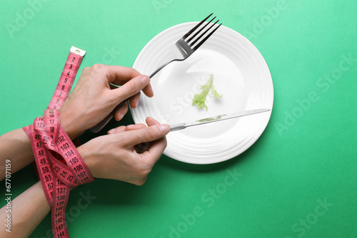 Diet concept. Woman holding cutlery in hands tied with measuring tape over plate with lettuce leaf on green background, top view