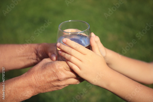 Child giving glass of water to elderly woman outdoors on sunny day, closeup