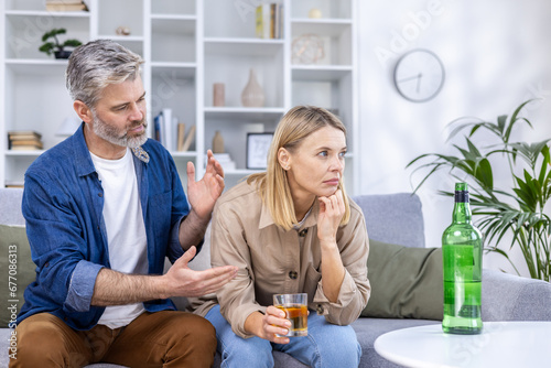 Billede på lærred Upset couple sitting in living room on sofa, alcoholic woman drinking a lot of strong alcohol alone, man trying to stop her, discuss everything, relationship problems, alcohol addiction
