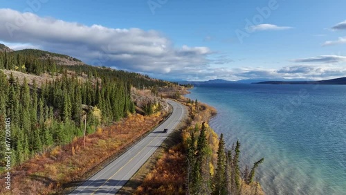 Automobile driving along highway in northern Yukon, Canada. Drone shot following the pick up truck as the road curves along the edge of a lake on a sunny day