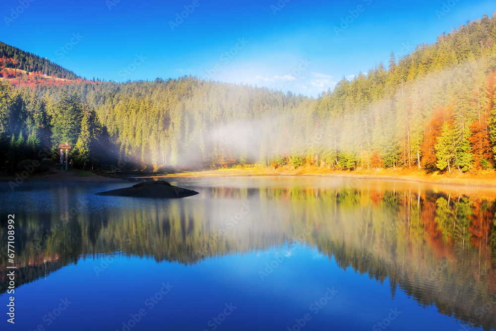 sunny landscape with lake in autumn. fog above the water surface. colorful scenery with sky and trees reflection
