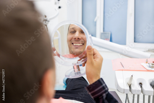 Happy Handsome Caucasian Man Looking To Mirror After Teeth Treatment In Modern Clinic, Male Patient Sitting In Chair In Stomatological Cabinet And Enjoying His New Smile