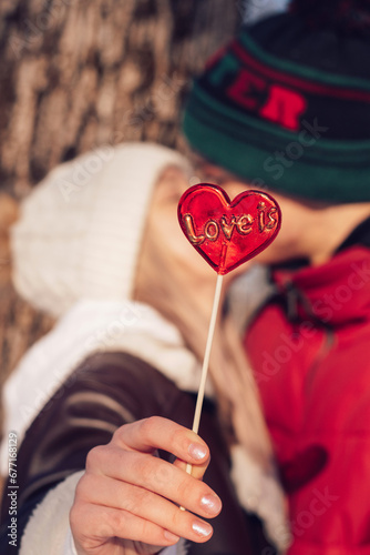 Blurred young couple in love kissing on a date on a winter day.In the foreground is a red lollipop in the shape of a heart and the inscription Love is.Valentine's day concept.