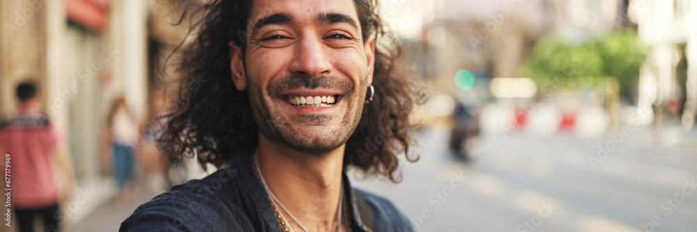 Obraz premium Young italian guy with long curly hair and stubble takes selfie on mobile phone while standing on the street. Stylish man posing smiling at camera of his phone
