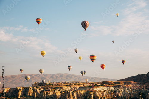 Hot air balloons ascend over the unique rock formations and valleys of Cappadocia, framed by a soft clouded sky