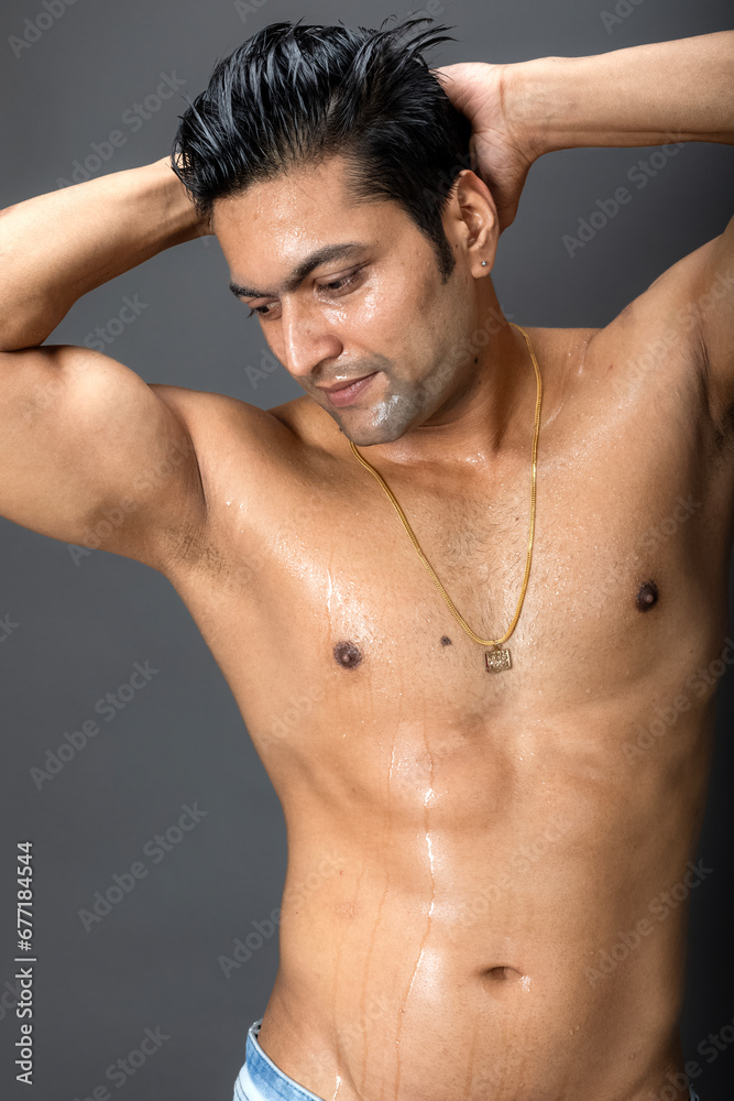 Portrait of a shirtless young man. Healthy muscular man with black hair, posing with hands behind head on grey background.