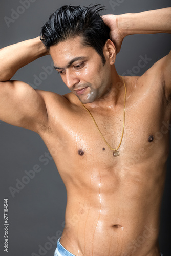 Portrait of a shirtless young man. Healthy muscular man with black hair, posing with hands behind head on grey background.