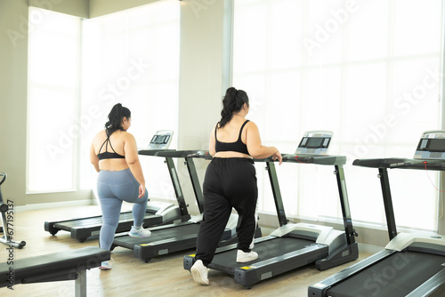 Two Asian overweight women running on treadmill in fitness club, healthy chubby female wearing sportswear exercising, stretching, and walking in gym, happy curvy girls in weight loss workout program photo