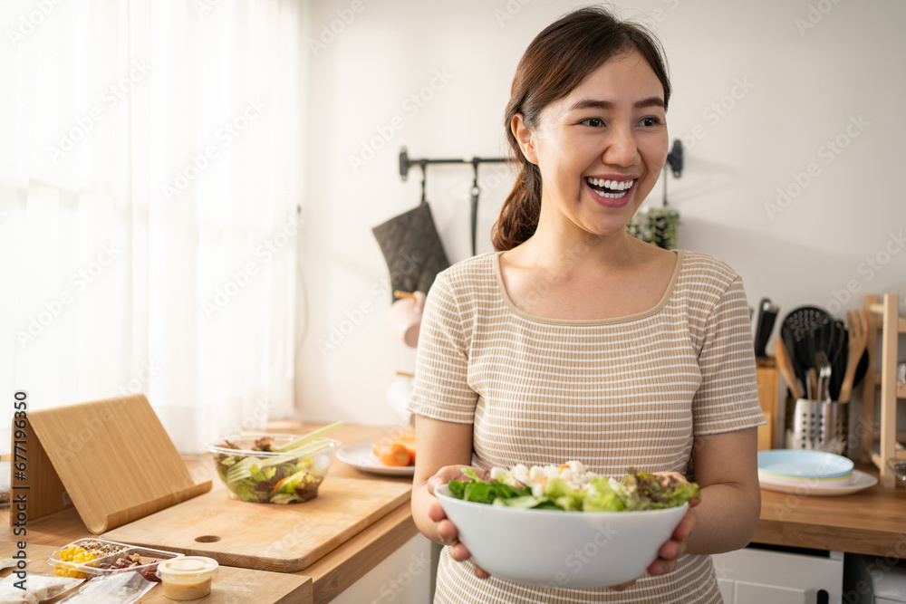 Happy smiling young Asian woman is preparing a fresh healthy vegan salad with many vegetables in the kitchen at home and trying a new recipe.