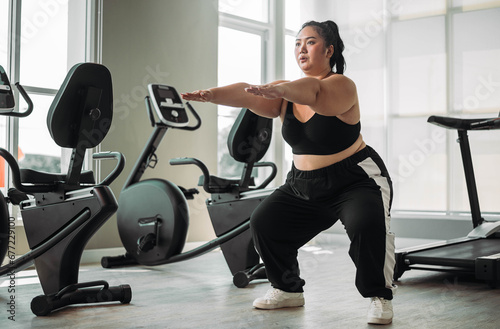Plus size Asian woman exercises in gym. Beautiful overweight woman in sportswear doing squat exercise in fitness. concept of body positive, self-acceptance, weight loss. photo