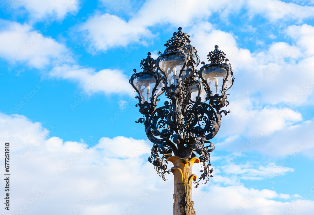 Street lamp against the blue sky with clouds . Lamp base features elaborate scrolls and curves, characteristic of the Baroque period