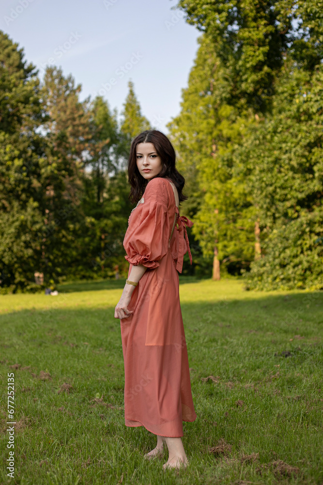 Portrait of a beautiful brunette girl in dress posing in the park, green tree background.
