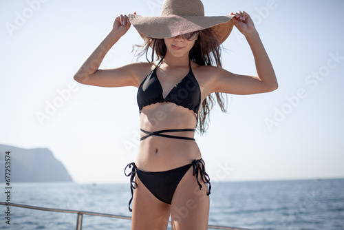 Photographie A beautiful young girl with a slender figure travels on a boat during a summer vacation in the open sea or ocean A model is wearing a swimsuit, sunglasses,straw hat