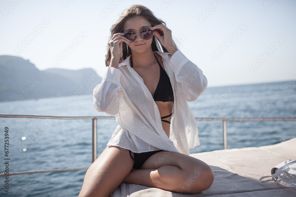 A beautiful young girl with a slender figure travels and rests on a boat during a summer vacation in the open sea or ocean . A model is wearing a black swimsuit, white shirt and sunglasses