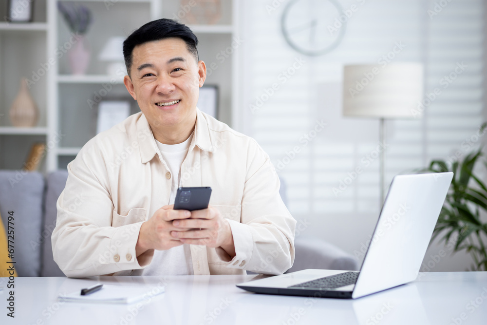 A happy Korean man is sitting in a bright living room at a table in front of a laptop, holding a phone in his hands, tapping on the screen, texting, smiling and looking at the camera. A happy Korean man is sitting in a bright living room at a table in front of a laptop, holding a phone in his hands, tapping on the screen, texting, smiling and looking at the camera.