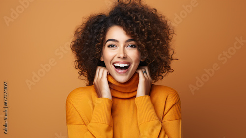 A joyful, beaming female adorned with cosmetics gleefully chuckling and signaling at the lens on an orange backdrop.