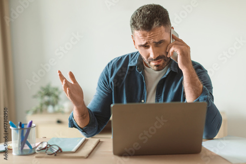 Discontented middle aged guy talking by cellphone at work desk
