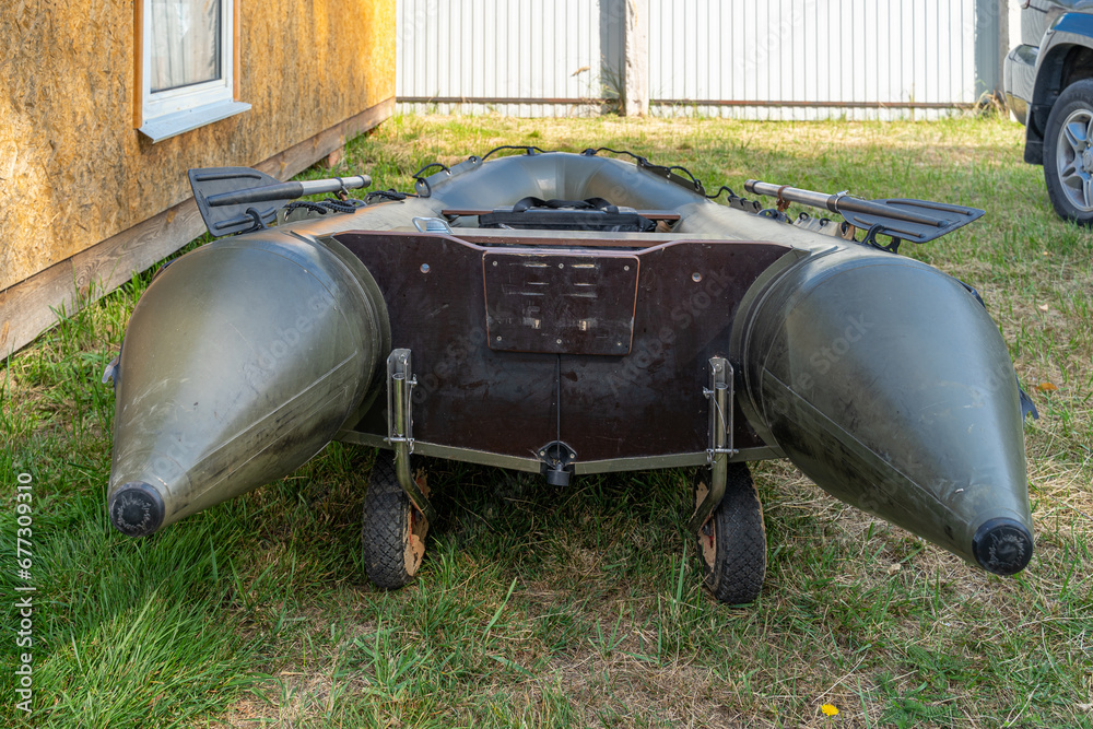 Green inflatable 4 seater boat with transom for motor and paddle for fishing and traveling with folding wheels for coastal mobility. The boat is lying on the grass on the shore after swimming. Green inflatable 4 seater boat with transom for motor and paddle for fishing and traveling with folding wheels for coastal mobility. The boat is lying on the grass on the shore after swimming.