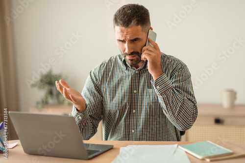 stressed man talking on cellphone managing tasks on laptop indoor