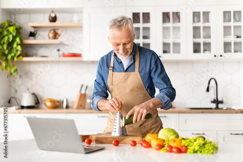 Senior Man In Apron Watching Video Tutorials While Cooking Food In Kitchen