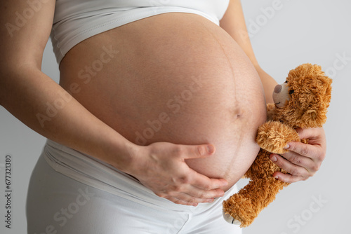 Close-up of a cuddly toy baer held by the mother's hand, who is looking excitedly at her naked, round belly and expecting the baby. Last month of pregnancy - week 39. White background. Bright shot. photo