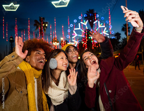 Group of young multiethnic friends in winter clothes making a selfie or video call during the night at a Christmas fair
