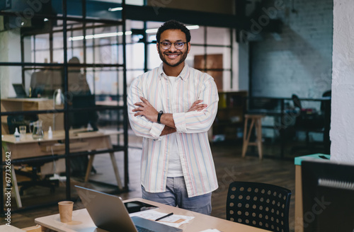Wallpaper Mural Cheerful ethnic man standing with folded hand by table with laptop in office Torontodigital.ca
