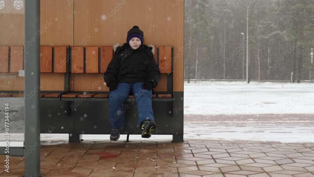Lonely sad child sits on bench outside on winter snowy day. Boy dangles his legs and looks at the snowfall, handheld shot. Loneliness and conflicts in the family.
