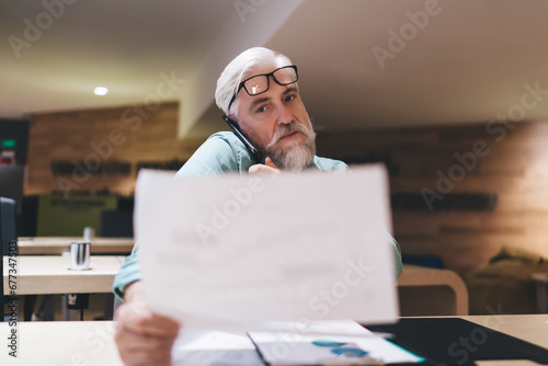 Senior Caucasian man with a beard, wearing glasses and a headset, looks pensively at a document in a modern office space, suggesting focused work and communication