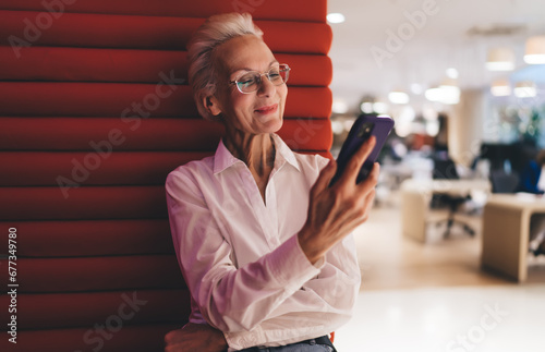 Contemplative senior Caucasian female professional reading on smartphone, sitting on red modern furniture, office background, reflecting