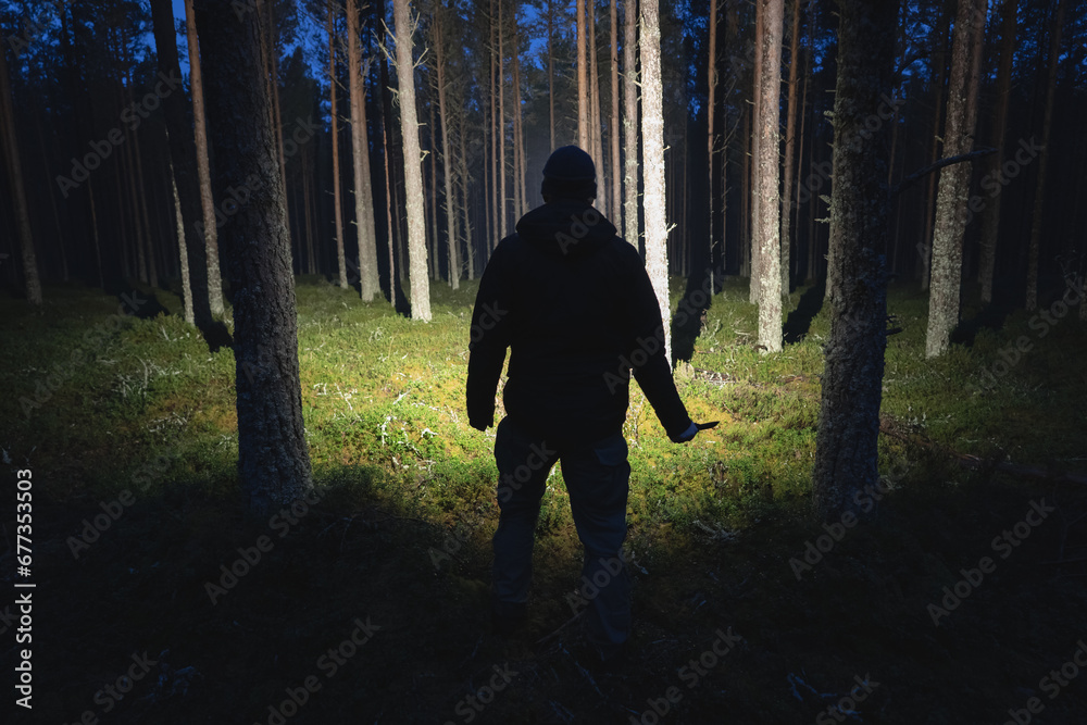 Silhouette of a man with a knife and a flashlight in a dark forest. - obrazy, fototapety, plakaty 