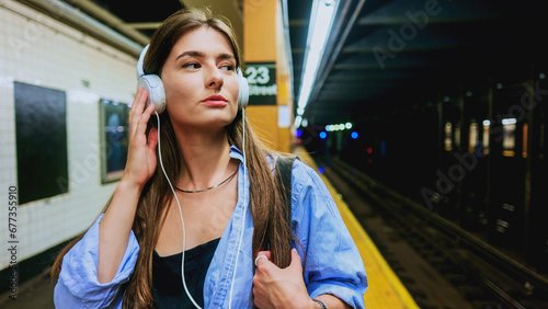 Young charming woman using mobile phone and listening music playlist with wireless headphones while waiting on tram station. City lifestyle concept. Travel in public transport. Female journey.