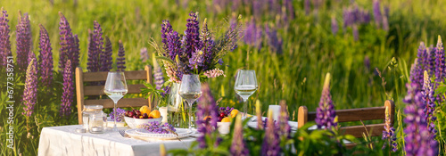 Elegant gorgeous wedding table decor or romantic dinner arrangement outdoors in blooming field. Purple lupine flowers, candles, fruits and wine, wooden furniture. Sunset, summer, golden hour banner
