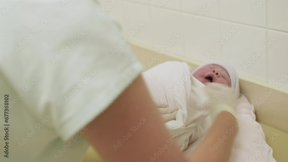Nurse in rubber gloves wraps newborn baby on changing table in hospital room. Child undergoes professional doctor movements but cries Nurse in rubber gloves wraps newborn baby on changing table in hospital room. Child undergoes professional doctor movements but cries