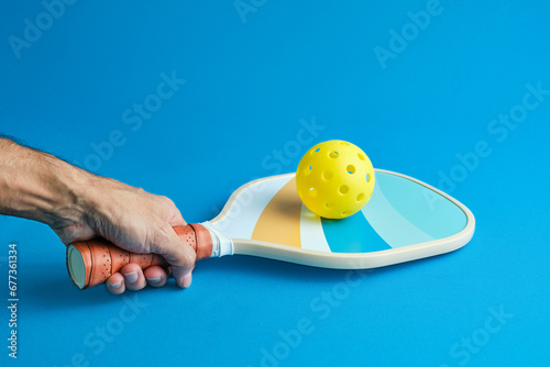 man's hand holding a pickleball paddle with a yellow ball on top on blue background