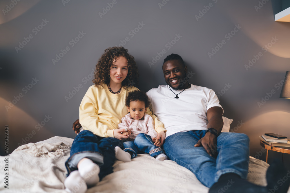 Happy multiracial family sitting together on cozy bed while looking at camera