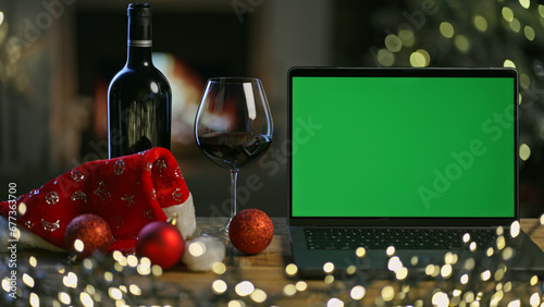 Glass and bottle of red wine with laptop computer on table in Christmas decorated room. Christmas tree and lights in winter home with fireplace. Green screen for web shop ads and vinery holiday offer.
