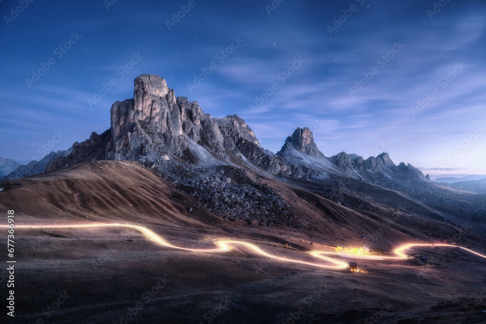 Car light trails on mountain road and high rocks at night in autumn in Passo Giau, Dolomites, Italy. Landscape with blurred light trails, hills, mountain peaks, blue sky with stars in fall at sunset