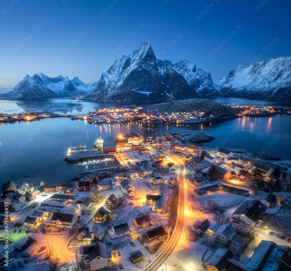 Aerial view of snowy village, islands, rorbu, city lights, blue sea, rocks and mountain at night in winter. Beautiful landscape with town, street illumination. Top view. Reine, Lofoten islands, Norway - obrazy, fototapety, plakaty 