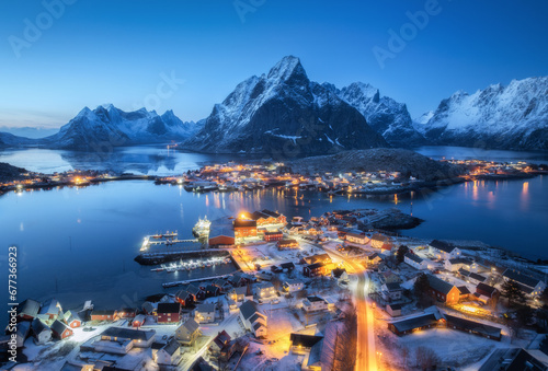 Fototapeta Aerial view of snowy village, islands, rorbu, city lights, blue sea, rocks and mountain at night in winter