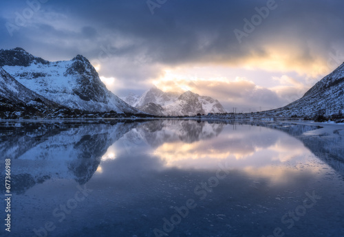 Beautiful snowy mountains and colorful sky with clouds and golden sunlight at sunset in winter in Lofoten islands, Norway. Landscape with rocks in snow, sea coast, reflection in water, overcast sky