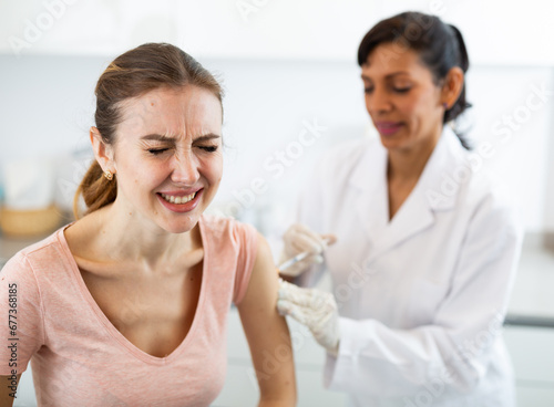 Latin woman physician making injection of covid-19 vaccine for caucasian woman writhing in pain.