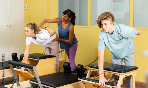 Young boy and girl doing exercises on pilates reformers. Their trainer hispanic woman correcting them.