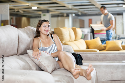 In furniture store,young girl is sitting on sofa with ottoman.She presents chosen sofa in her cozy apartment