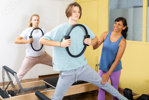Young boy and girl doing exercises on pilates reformers and using special rings. Their trainer hispanic woman correcting them.
