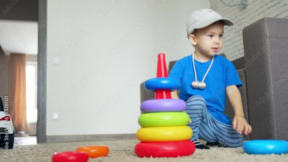 Smiling Caucasian baby boy comes up to a toy pyramid. Kid in a cap sits down on the floor to play with his toy. Low angle view.