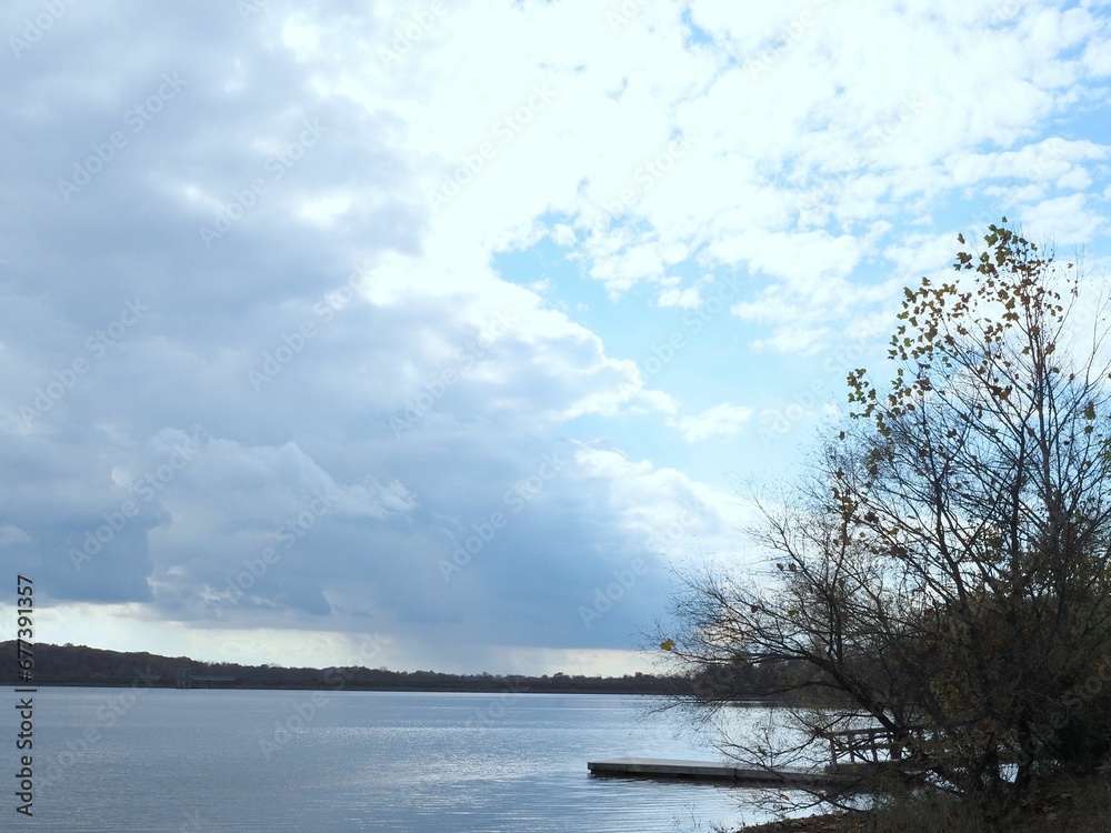 Cloudy sky above the smooth lake with leafless trees on the shore.